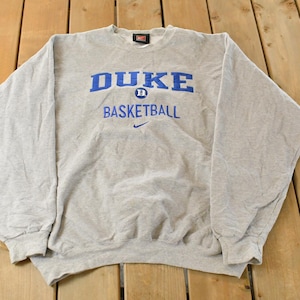 Może przedstawiać: Jasnoszara bluza z napisem "DUKE BASKETBALL" w niebieskim kolorze. Logo Nike jest widoczne pod tekstem. Bluza ma długie rękawy i okrągły dekolt.