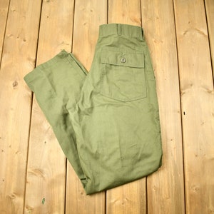 May include: A pair of olive green trousers with a single back pocket. The trousers are folded in half and laid flat on a wooden surface.
