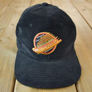 May include: Black corduroy baseball cap with an embroidered logo. The logo features a circular design with the word "CANUCKS" and a stylized graphic in orange, yellow, and white.