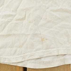 May include: Close-up of a white, slightly wrinkled fabric with several brown stains. The bottom edge of the fabric is visible, showing a hem. The fabric appears to be a t-shirt or similar garment.