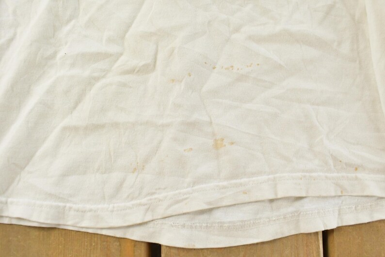 May include: Close-up of a white, slightly wrinkled fabric with several brown stains. The bottom edge of the fabric is visible, showing a hem. The fabric appears to be a t-shirt or similar garment.