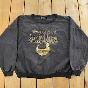 Puede incluir: Una sudadera negra con el texto dorado "Lifestyles of the Poor and Aimless of Lake Vermilion". La sudadera tiene cuello redondo, mangas largas con puños negros y dobladillo negro. El diseño incluye un diseño circular.