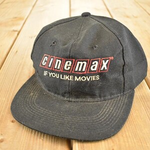 Könnte beinhalten: Schwarze Baseballkappe mit dem Cinemax-Logo in Rot und Weiß und dem Text "IF YOU LIKE MOVIES" darunter. Die Kappe hat einen gebogenen Schirm und einen Knopf oben. Auf einer Holzoberfläche.