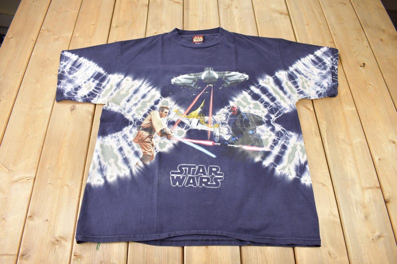 May include: A blue tie-dye t-shirt with a Star Wars graphic featuring characters from the movie. The graphic shows a lightsaber duel between a Jedi and a Sith Lord. The text "STAR WARS" is printed below the graphic.