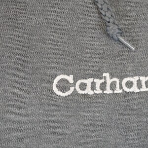 Puede incluir: Primer plano de una sudadera con capucha gris con el nombre de la marca "Carhartt" bordado en blanco. El cord&oacute;n gris se extiende desde la parte superior. La tela tiene una apariencia texturizada.