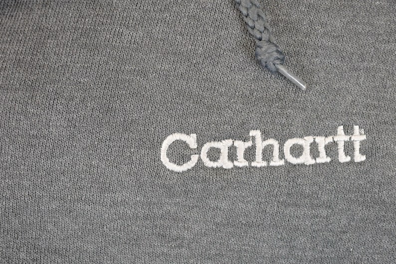 Puede incluir: Primer plano de una sudadera con capucha gris con el nombre de la marca "Carhartt" bordado en blanco. El cord&oacute;n gris se extiende desde la parte superior. La tela tiene una apariencia texturizada.