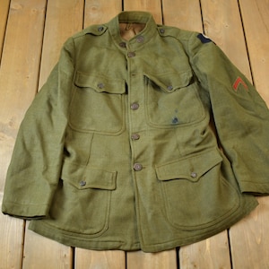 May include: A green military jacket with a high collar and multiple pockets. The jacket has a button closure and a patch on the sleeve.