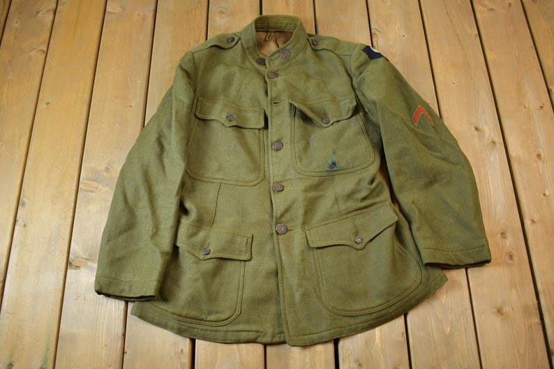 May include: A green military jacket with a high collar and multiple pockets. The jacket has a button closure and a patch on the sleeve.