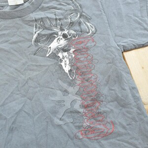 May include: Gray t-shirt featuring a graphic of a deer skull with antlers and a red, stylized design. The shirt has a crew neck and short sleeves. The design is printed on the front of the shirt.