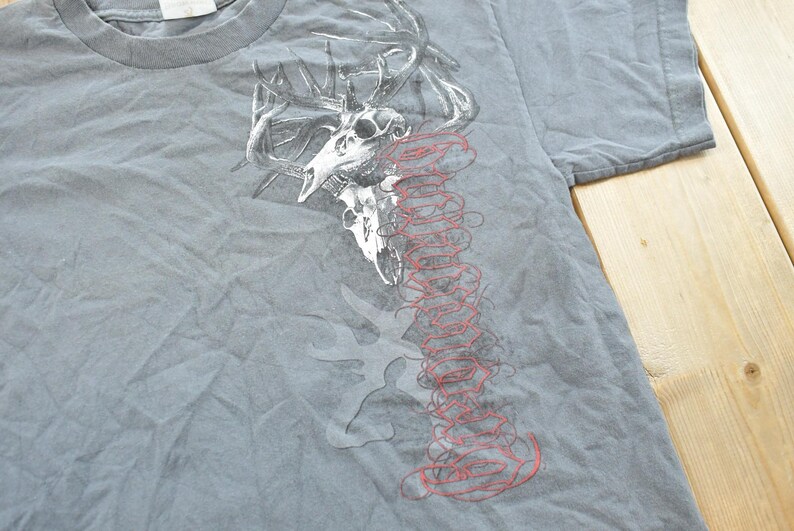May include: Gray t-shirt featuring a graphic of a deer skull with antlers and a red, stylized design. The shirt has a crew neck and short sleeves. The design is printed on the front of the shirt.