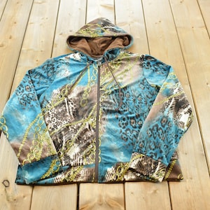 Puede incluir: Una sudadera con capucha y cremallera que presenta un vibrante estampado animal en tonos turquesa, marrón y negro, superpuesto con un diseño de cadenas doradas. La prenda incluye una capucha con forro marrón, mangas largas y cierre de cremallera.