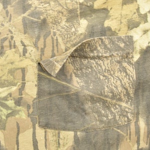May include: Close-up of a camouflage pattern fabric with a pocket. The fabric features a mix of brown, green, and tan colors, creating a woodland camouflage design. The pocket is rectangular and sewn onto the fabric.