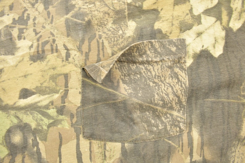 May include: Close-up of a camouflage pattern fabric with a pocket. The fabric features a mix of brown, green, and tan colors, creating a woodland camouflage design. The pocket is rectangular and sewn onto the fabric.