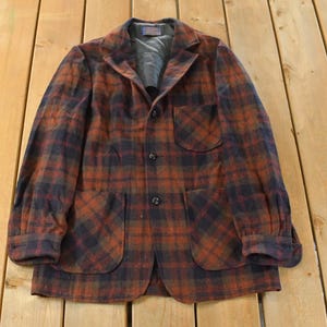 May include: A vintage plaid blazer in shades of rust, navy, and black. The jacket has a notched collar, two front pockets, and a three-button closure. The sleeves are cuffed, and the blazer is laid flat on a wooden surface.