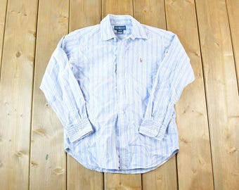 May include: A light blue and white striped button-down shirt. The long-sleeved shirt has a classic collar and a small embroidered logo on the chest. The shirt is made of a lightweight fabric and is laid flat on a wooden surface.