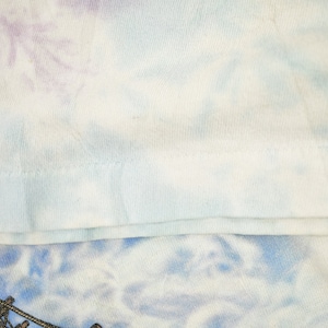 May include: Close-up of a light blue tie-dye t-shirt with a white seam along the bottom edge.