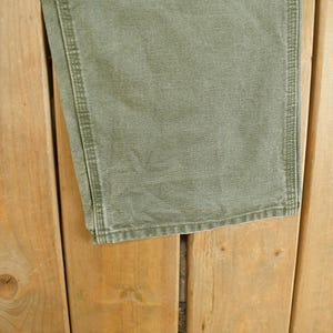 Puede incluir: Primer plano de unos pantalones verde oliva con costuras visibles a lo largo de las costuras. Los pantalones est&aacute;n hechos de una tela duradera y parecen ligeramente desgastados. Los pantalones est&aacute;n sobre una superficie de madera.