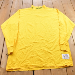 May include: A yellow long-sleeved shirt with a mock neck collar. The shirt has the word "PACKERS" embroidered on the collar in green and black lettering. The shirt has a white tag with the words "MAXIT" and "WORLD CHAMPIONSHIP" printed on it.