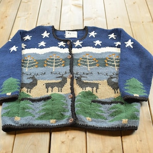 May include: A long-sleeve button-down knit cardigan featuring a dark blue background with a white star pattern at the top. The midsection displays a landscape with deer and trees in shades of tan, blue, and green, with two front pockets.
