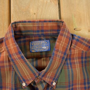 May include: Close-up of a button-down shirt with a plaid pattern in shades of orange, green, and blue. The shirt collar is visible, along with a blue label that reads "Pendleton". The shirt is made of a soft, woven fabric.