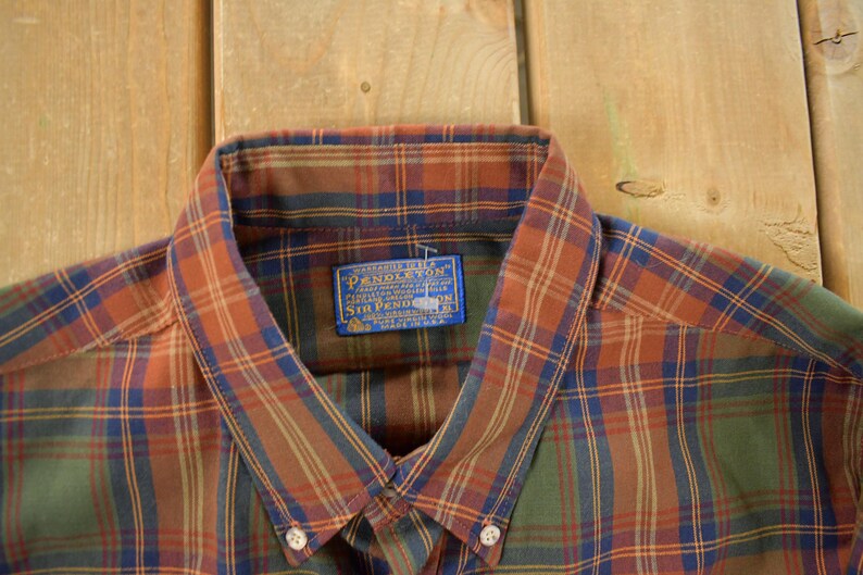 May include: Close-up of a button-down shirt with a plaid pattern in shades of orange, green, and blue. The shirt collar is visible, along with a blue label that reads "Pendleton". The shirt is made of a soft, woven fabric.