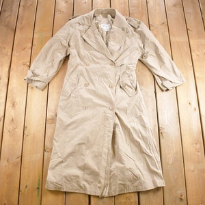 Könnte beinhalten: Ein beiger Trenchcoat mit doppelreihigem Verschluss und Gürtel. Der Mantel hat zwei Taschen vorne und eine lange, fließende Silhouette.