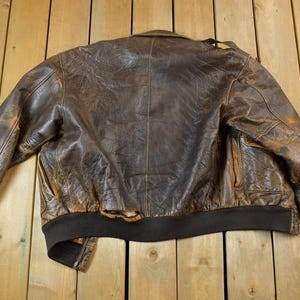 May include: A vintage brown leather bomber jacket with a distressed appearance. The jacket features a black ribbed collar, cuffs, and hem. The leather shows wear and tear, adding to its classic style.