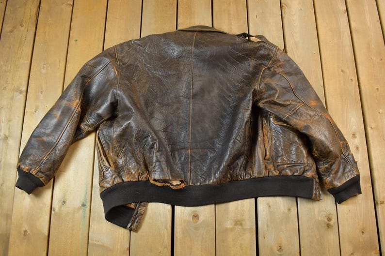 May include: A vintage brown leather bomber jacket with a distressed appearance. The jacket features a black ribbed collar, cuffs, and hem. The leather shows wear and tear, adding to its classic style.