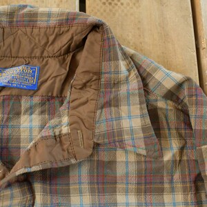 Puede incluir: Primer plano de una camisa a cuadros con botones y cuello acolchado marr&oacute;n. La camisa tiene una etiqueta azul con texto blanco. El estampado a cuadros incluye l&iacute;neas beige, azules y rojas.