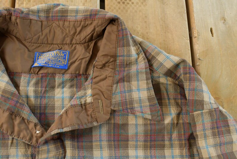 Puede incluir: Primer plano de una camisa a cuadros con botones y cuello acolchado marr&oacute;n. La camisa tiene una etiqueta azul con texto blanco. El estampado a cuadros incluye l&iacute;neas beige, azules y rojas.