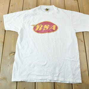 Könnte beinhalten: Weißes T-Shirt mit einem roten und gelben Logo, auf dem "BSA" steht.