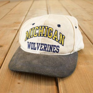 May include: A vintage baseball cap with a cream-coloured crown and a grey brim. The cap features the text "MICHIGAN WOLVERINES" in yellow and navy blue embroidery, a classic sports-themed accessory.