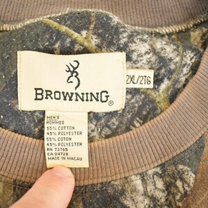 May include: Close-up of a camouflage-patterned Browning sweatshirt. The tag reads "BROWNING" with a deer logo, and includes fabric composition details. The sweatshirt has a brown neckline and the size is 2XL/2TG.