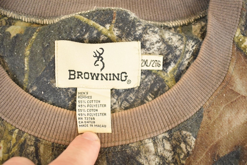 May include: Close-up of a camouflage-patterned Browning sweatshirt. The tag reads "BROWNING" with a deer logo, and includes fabric composition details. The sweatshirt has a brown neckline and the size is 2XL/2TG.
