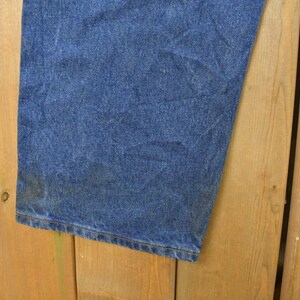 K&ouml;nnte beinhalten: Nahaufnahme eines Abschnitts blauer Jeans. Der Stoff weist Abnutzungserscheinungen und Verf&auml;rbungen in der N&auml;he des Saums auf. Die Jeans sind vor einem Holzgrund platziert.