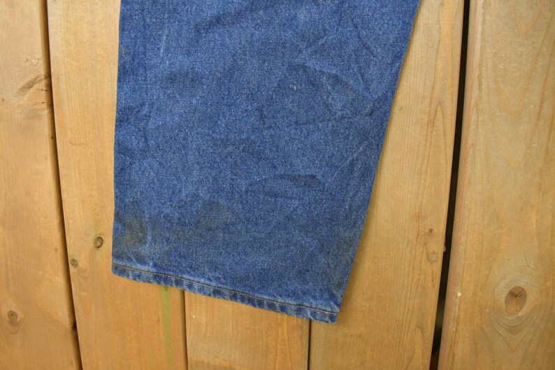 K&ouml;nnte beinhalten: Nahaufnahme eines Abschnitts blauer Jeans. Der Stoff weist Abnutzungserscheinungen und Verf&auml;rbungen in der N&auml;he des Saums auf. Die Jeans sind vor einem Holzgrund platziert.