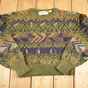 Könnte beinhalten: Ein Vintage-Pullover mit Rundhalsausschnitt und geometrischem Muster in Grün-, Lila-, Braun- und Schwarztönen. Der Pullover hat einen gerippten Kragen, Bündchen und Saum. Der Markenname "HARNESS" ist auf einem Etikett am Kragen zu sehen.