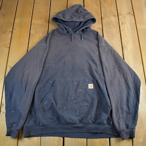 K&ouml;nnte beinhalten: Ein marineblauer Kapuzenpullover mit K&auml;ngurutasche und kleinem Logo-Patch. Der Sweatshirt hat Farbspritzer und liegt auf einer Holzoberfl&auml;che. Das Carhartt-Logo ist auf der Vordertasche zu sehen.