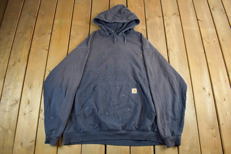 K&ouml;nnte beinhalten: Ein marineblauer Kapuzenpullover mit K&auml;ngurutasche und kleinem Logo-Patch. Der Sweatshirt hat Farbspritzer und liegt auf einer Holzoberfl&auml;che. Das Carhartt-Logo ist auf der Vordertasche zu sehen.