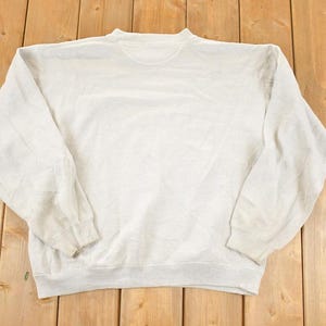 K&ouml;nnte beinhalten: Ein hellgraues Sweatshirt mit Rundhalsausschnitt und langen &Auml;rmeln. Das Sweatshirt besteht aus einem weichen Material und hat ein leicht abgenutztes Aussehen. Das Kleidungsst&uuml;ck liegt flach auf einer Holzoberfl&auml;che.