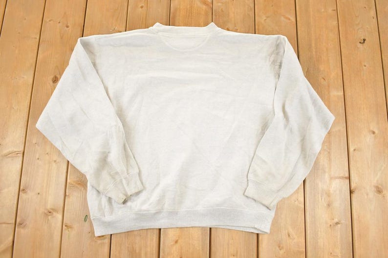 K&ouml;nnte beinhalten: Ein hellgraues Sweatshirt mit Rundhalsausschnitt und langen &Auml;rmeln. Das Sweatshirt besteht aus einem weichen Material und hat ein leicht abgenutztes Aussehen. Das Kleidungsst&uuml;ck liegt flach auf einer Holzoberfl&auml;che.
