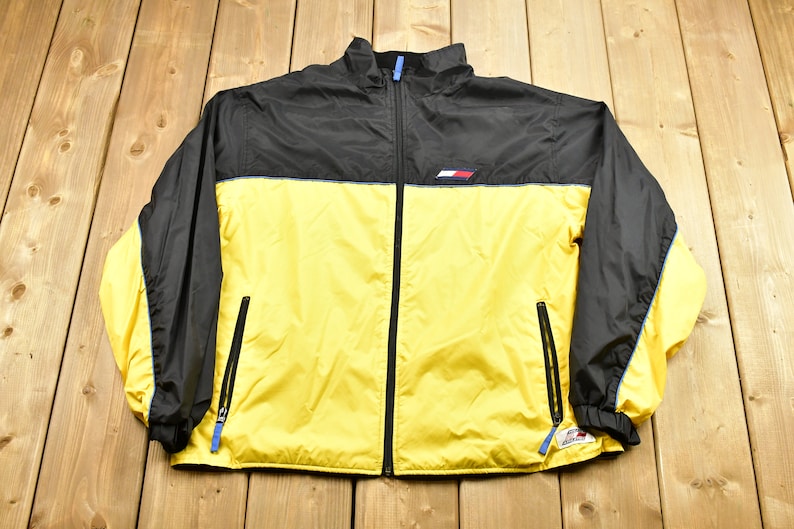 Pu&ograve; includere: Una giacca a vento nera e gialla con una cerniera blu e un piccolo logo sul petto.