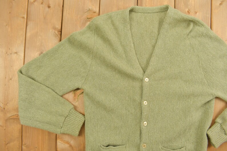 May include: A light green knitted cardigan with a V-neck and button closure. The cardigan has long sleeves and two pockets on the front.