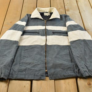 May include: A slate blue and off-white color-blocked jacket featuring a full-length front zipper, a fold-down collar, and two horizontal zippered chest pockets. The garment has long sleeves with elasticized cuffs and a straight hemline design.