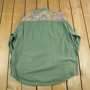 May include: Green long-sleeve shirt with a camouflage pattern on the upper back. The shirt has a button-down collar and a curved hem. The fabric appears to be a durable, possibly workwear-style material.