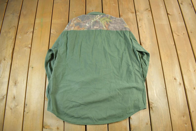 May include: Green long-sleeve shirt with a camouflage pattern on the upper back. The shirt has a button-down collar and a curved hem. The fabric appears to be a durable, possibly workwear-style material.