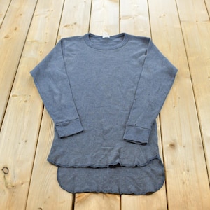 May include: A long-sleeve charcoal gray thermal shirt featuring a crew neckline, ribbed cuffs, and a curved high-low hemline. The garment is constructed from a textured waffle-knit fabric and includes a small white tag at the inner collar.