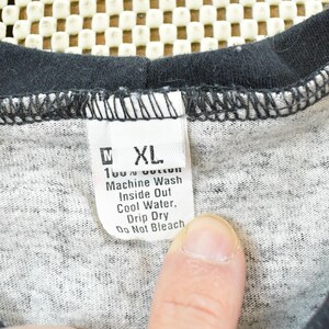 May include: Close-up of a gray and black t-shirt with a tag. The tag reads "XL" and washing instructions: "Machine Wash Inside Out Cool Water, Drip Dry Do Not Bleach". The shirt is made of 100% cotton.