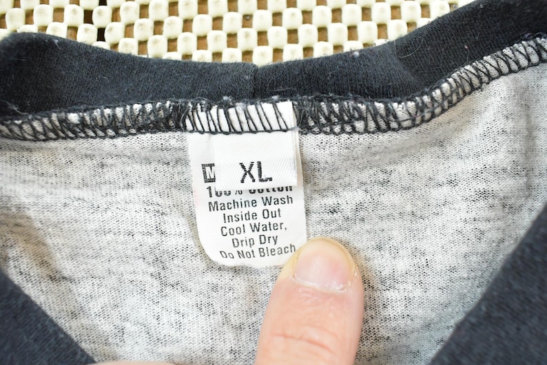 May include: Close-up of a gray and black t-shirt with a tag. The tag reads "XL" and washing instructions: "Machine Wash Inside Out Cool Water, Drip Dry Do Not Bleach". The shirt is made of 100% cotton.