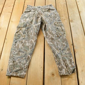 May include: Camouflage cargo pants with multiple pockets, in shades of brown, green, and gray. The pants are laid flat on a wooden surface, showcasing the detailed pattern and design.